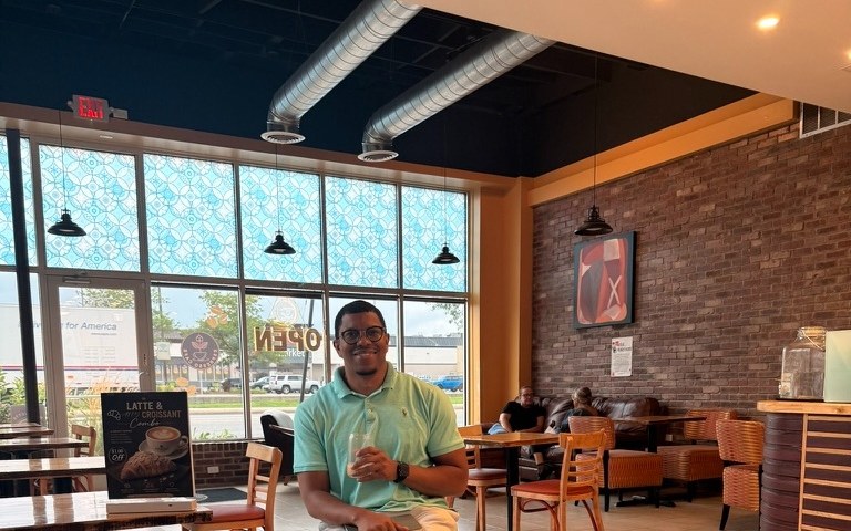 Takkeem Morgan sitting in Espresso Cafe, the open room with warm pendant lights, brick wall, and patterned window film visible around him